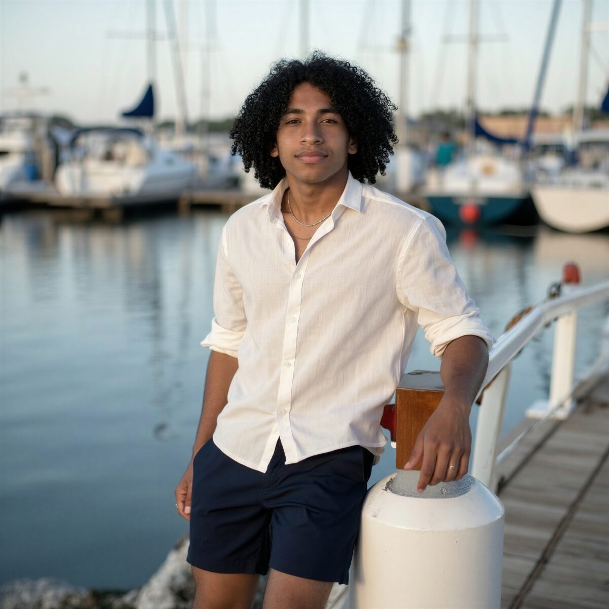 Man in white shirt and navy shorts posing confidently by marina with yachts