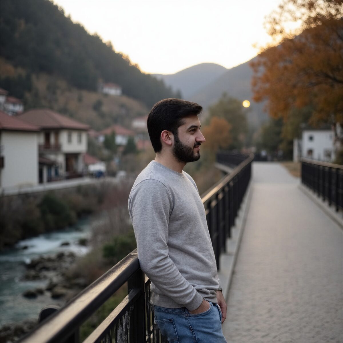 Man in grey sweater leaning on a bridge railing with scenic mountains and river at sunset